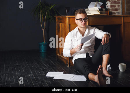 Stattliche Schriftsteller Mann mit Brille und weißem T-Shirt sitzen auf dem Boden in der Nähe des Tisches mit Schreibmaschine, Kaffee trinken und lesen Neues Kapitel seines Buches Stockfoto