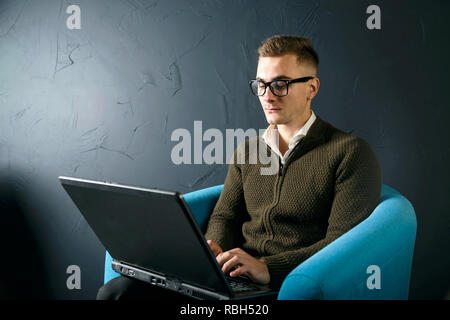 Stattliche ernsthaften jungen Mann mit grünen Pullover sitzen auf die türkisfarbene Sessel und mit Laptop Stockfoto