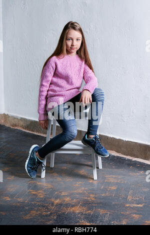 Zuversichtlich Kind mädchen mode Pose sitzt auf einem Stuhl, konkrete weißen Hintergrund. Elegantes Modell in rosa Pullover. Schöne glamourösen Kind Jugendlicher in Kleidung casual, urbanen Stil. Studio gedreht. Stockfoto