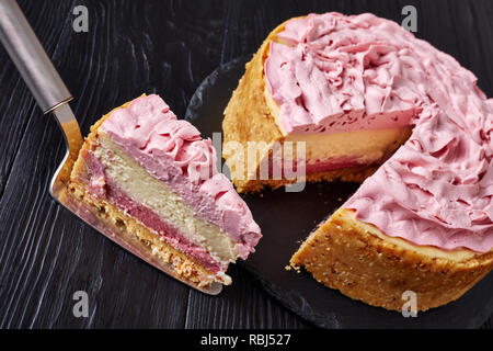 White Chocolate Raspberry cheesecake gekrönt mit Frischkäse gefärbt mit Boden Himbeeren auf einen schwarzen Stein Fach auf Holztisch, in Scheiben geschnitten, Ansicht von Stockfoto