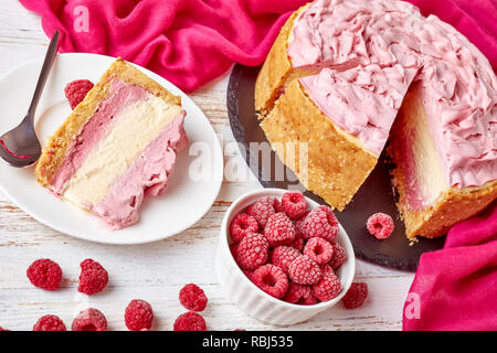 Teil einer White Chocolate Raspberry cheesecake auf einer Platte und geschnittene Käsekuchen auf einen schwarzen Stein Fach mit Stoff und gefrorene Himbeeren auf einer weißen Stockfoto