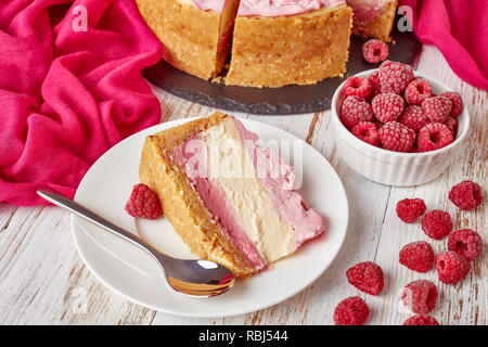 Nahaufnahme eines Teils einer geschichteten Raspberry cheesecake auf einem Teller. geschnitten Käsekuchen auf einen schwarzen Stein Fach mit Stoff und gefrorene Himbeeren auf einer whi Stockfoto