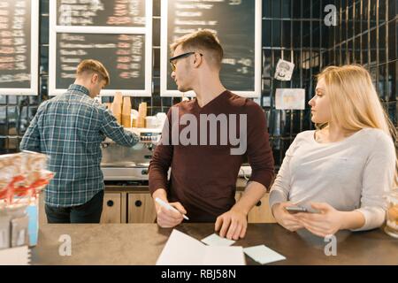 Die jungen Menschen arbeiten im Coffee Shop, Mann und Frau in der Nähe von Bar, Empfang per Telefon, Barista auf dem Hintergrund der Kaffeemaschine. Teamwork, Stockfoto