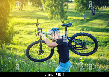 Radfahrer Mann in Helm trägt sein Mountainbike im Aufstieg im Hintergrund grüne Natur. Konzept der aktiven und gesunden Lebensstil Stockfoto