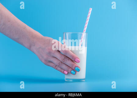 Die Hand einer Frau hält ein Glas frische Milch mit einem Strohhalm auf blauem Hintergrund. Bunte Minimalismus. Das Konzept der gesunde Milchprodukte mit Kalzium Stockfoto