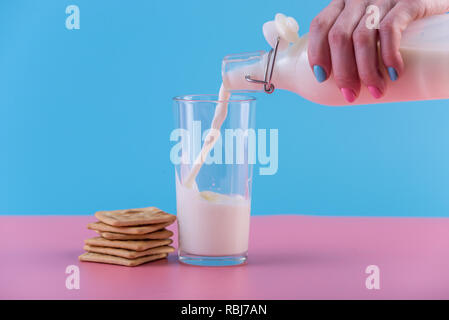 Die Hand einer Frau giesst frische Milch aus einem Glas Flasche in ein Glas und Cookies auf ein Pastell Hintergrund. Bunte Minimalismus. Das Konzept der gesunden Milchprodukten Stockfoto