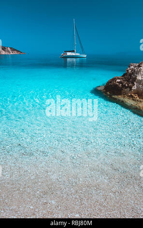 Einsamen weißen Segeln Katamaran drift auf ruhigen Meer Oberfläche. Reine Azure sauber blaue Lagune mit flachem Wasser und Pebble Beach. Einige braune rock Steine im Vordergrund Stockfoto