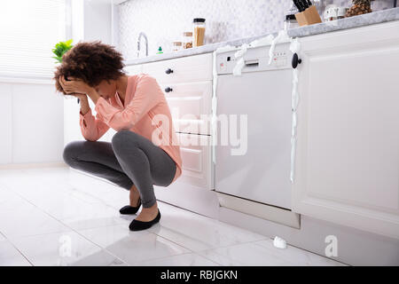 Sorgen junge Frau sitzt Vor der beschädigten Geschirrspüler mit Schaum Aus Aus Stockfoto