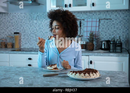 Junge afrikanische Frau Essen ein Stück Kuchen mit Löffel in der Küche sitzt Stockfoto