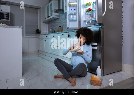 Junge schwangere Frau Essen Stück Kuchen sitzen vor dem offenen Kühlschrank in der Küche Stockfoto