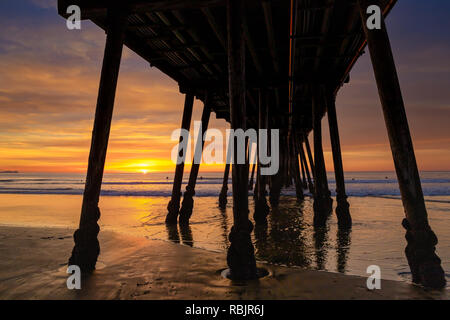 Die untergehende Sonne am Pier in Imperial Beach, Kalifornien mit den Silhouetten der Surfer warten auf Wellen. Stockfoto