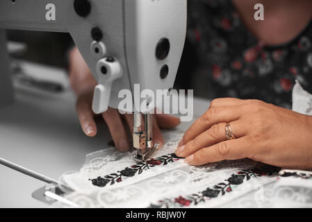 Nahaufnahme von Frau Näherin Hände verbinden weißen Stoff details mit Blumen bunte Stickerei mit langen Streifen der Spitze auf moderne Nähmaschine. Stockfoto