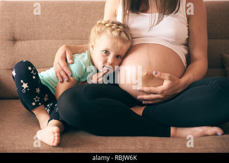 Schöne Mädchen sitzt auf einem Sofa mit schwangeren Mutter, Kamera schaut. Fröhliche Frau, Tochter und erwarten Baby. Konzept der elterlichen Liebe ein Stockfoto
