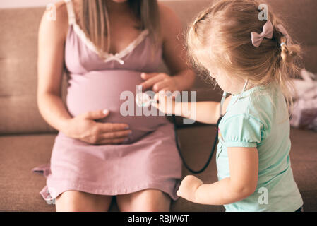 Selektiver Fokus der niedlichen Tochter spielen Arzt und Prüfung der schwangeren Mutter. Glückliche Frau auf dem Sofa zu Hause sitzen und spielen mit schöne Mädchen. Anhand von quantitativen Simulatio Stockfoto