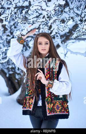 Junge schöne Langhaarige Frau in weiß bestickt Bluse und bunten Weste im Freien auf sonnigen Wintertag auf verschneiten Landschaft Hintergrund. Tradit Stockfoto