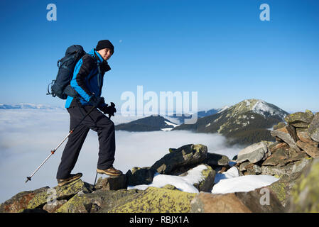Touristische Wanderer mit Rucksack und Wanderstöcke Klettern an steilen felsigen Berghang auf dem Hintergrund des blauen Himmels, neblige Tal mit weißen Clo gefüllt Stockfoto