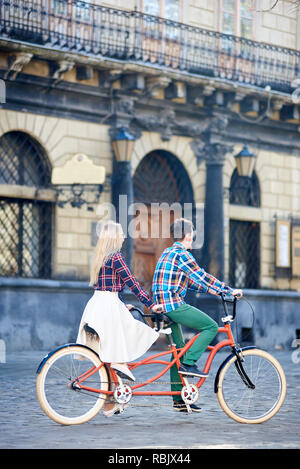 Rückansicht des Jungen gerne touristische Paar, bärtigen Mann und Langhaarige blonde Frau in weißen Rock, Tandem Bike entlang der befestigten Stadt Bürgersteig auf hellen Stockfoto