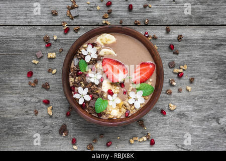 Schüssel mit Schokolade und Banane Smoothie mit Schokolade Müsli, Erdbeere und Granatapfel Samen mit Blumen dekoriert. top anzeigen. gesundes Frühstück Stockfoto