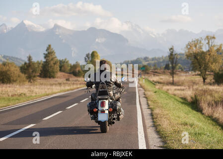 Rückansicht der Biker in schwarzem Leder Jacke reiten Motorrad entlang der Straße auf unscharfen Hintergrund der schönen Bergwelt mit schneebedeckten Gipfeln, Veh verschieben Stockfoto