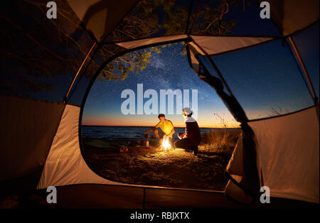 Camping am Meer bei Sonnenuntergang, Ansicht von innen touristische Hütte. Junge touristische Paar, Mann und Frau, die Zubereitung von Speisen auf Gas Brenner, sitzen in der Nähe von Lagerfeuer Stockfoto