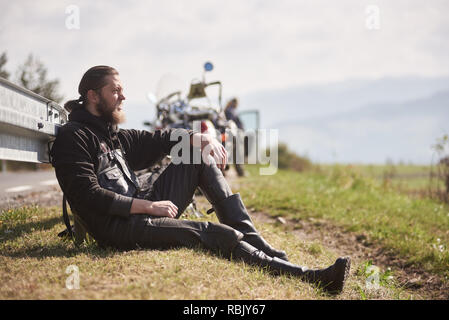 Athletisch sportliche Motorradfahrer in schwarzem Leder Bekleidung zu leistungsstarken Motorrad auf Gras am Straßenrand sitzen auf dem Hintergrund von Sunny die fernen Hügel unter strahlend blauem Himmel verschwimmt. Stockfoto