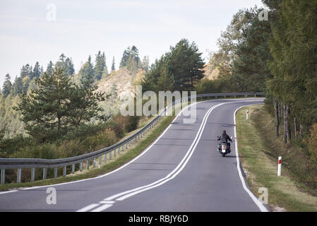 Rückansicht der Motorradfahrer in schwarzem Leder Outfit mit langen Haaren, Cruiser Motorrad twisted leer Asphaltstraße auf hellen sonnigen Sommertag durch grünen Wald Bäume unter klaren blauen wolkenlosen Himmel. Stockfoto
