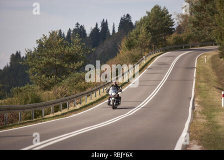 Motorradfahrer in schwarzem Leder outfit Reiten cruiser Motorrad entlang Twisted Straße auf die hellen sonnigen Sommertag auf dem Hintergrund der nebligen entfernte grün bewaldeten Hügeln unter blauen Morgenhimmel. Stockfoto