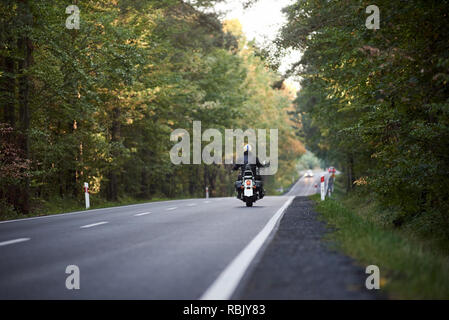 Rückansicht der Motorradfahrer in schwarzem Leder Jacke und weißen Helm reiten Cruiser Motorrad entlang der hügeligen Straße zwischen hohen grünen Bäumen. Aktiver Lebensstil, Liebe Abenteuer Konzept. Stockfoto