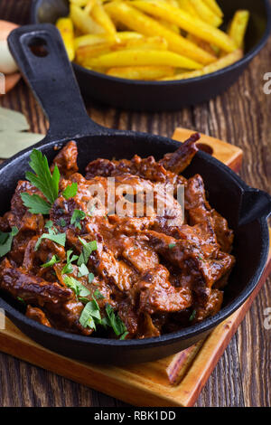 Stroganoff mit Tomatensauce und saure Sahne und gebratenen Kartoffeln in Gusseisen Skillet auf rustikalen Holztisch serviert. Stockfoto