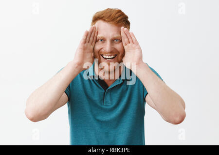 Studio shot der spielerische und freudige gut aussehende junge rothaarige Vater mit Borsten holding Palmen entlang Gesicht und breit grinsend, peekaboo Spielen mit Kindern und Spaß im Kreis der Familie über graue Wand Stockfoto
