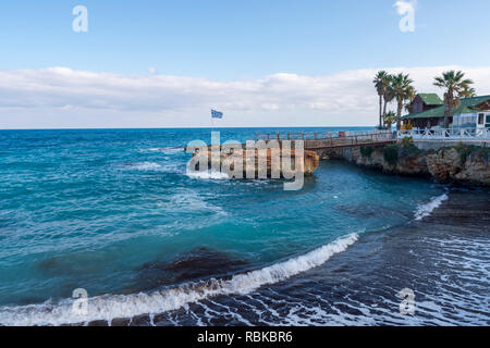 Bucht mit einem zurücktretenden Wave verlassen Schaum Streifen hinter, Cliff mit griechischer Flagge und Horizont und blauer Himmel im Hintergrund, Bild von in der Nähe der Kleinen Stockfoto