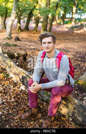 Jungen kaukasischen Mann mit Rucksack sitzen und entspannen auf den gefallenen Baum im herbstlichen Wald. weg schauen. lächelnden Kerl. Vertikale Karte. Selektive Schwerpunkte Stockfoto