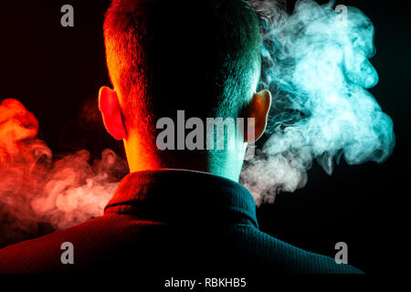 Ein Blick von hinten auf den Kopf von einem Kerl in einem T-Shirt Rauchen eine vape und Ausatmen mehrfarbige Rauch von Grün und Rot in verschiedenen Richtungen von ihm Stockfoto