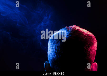 Ein Blick von hinten auf den Kopf eines Mannes in einem T-Shirt Rauchen eine vape und Ausatmen mehrfarbige Rauch von Blue in verschiedene Richtungen von sich selbst auf einem Stockfoto