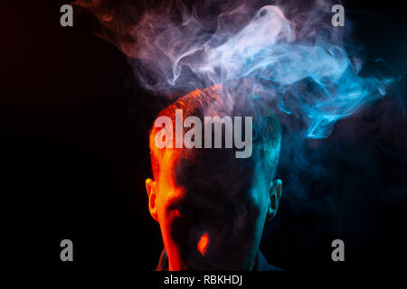 Das Gesicht eines Mannes im Schatten und Kopf ist rot, der raucht eine vape hervorgehoben und heraus gibt, mehrfarbige Rauch von Blau und Rosa über seinem Kopf, causi Stockfoto