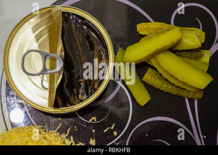 Ein Flach, oben ein schwarzes und weißes Glas Schneidebrett mit einer offenen Dose Sprotten in Öl, gehackte gesalzene grüne Gurken und geriebenem Käse Stockfoto