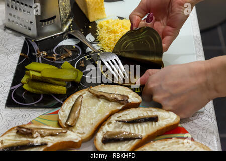 Blick auf die Hände des Mädchens, das Frühstück macht, indem Sie eine Dose Sprotten in Öl, auf denen liegt eine Gabel für Sandwiches aus Brot mit geriebenem Käse Stockfoto