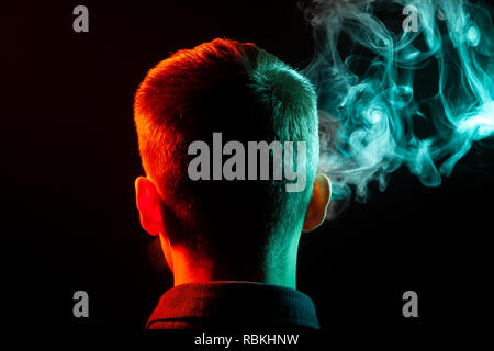 Ein Blick von hinten auf den Kopf eines Jungen in einem T-Shirt Rauchen eine vape und farbigen Ausatmen grünen Rauch in der rechten Richtung, aus sich selbst und mit der Hintergrundbeleuchtung Stockfoto