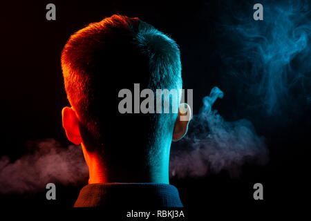 Ein Blick von hinten auf den Kopf eines Jungen in einem T-Shirt Rauchen eine vape und Ausatmen mehrfarbige Rauch von Grün und Rot in verschiedenen Richtungen von ihm Stockfoto