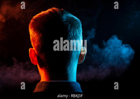 Ein Blick von hinten auf den Kopf von einem Kerl in einem T-Shirt Rauchen eine vape und Ausatmen mehrfarbige Rauch von Grün und Rot in verschiedenen Richtungen von ihm Stockfoto