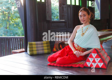 Junge schöne Thai asiatische Frau Dressing vintage Traditionelle Thai Kostüm sitzt auf Dreieck Kissen auf dem Balkon. Thailand kulturelle und traditionelle Tour Stockfoto