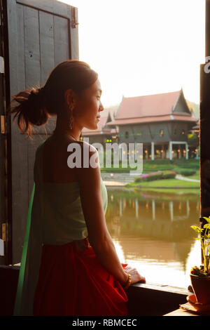 Junge schöne Thai asiatische Frau Dressing in Vintage Retro Traditionelle Thai Kostüm aus dem Fenster zu schauen. Thailand kulturellen und traditionellen Tourismus, Stockfoto