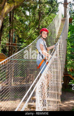 Teenager boy trug Gurt, Trageband und System belay Eingabe Hochseilgarten im Outdoor treetop Adventure Park durch hängende Strickleiter Stockfoto