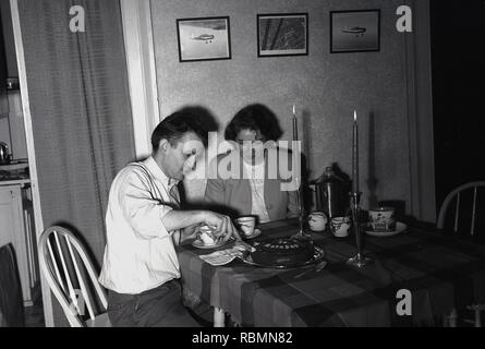 1950 eveningtime in eine kleine Wohnung und einen gemütlichen Geburtstag. Ein paar sitzen an kleinen Tuch bedeckt Tisch mit zwei Kerzen angezündet, als der Mann die Schokolade Kuchen. Stockfoto