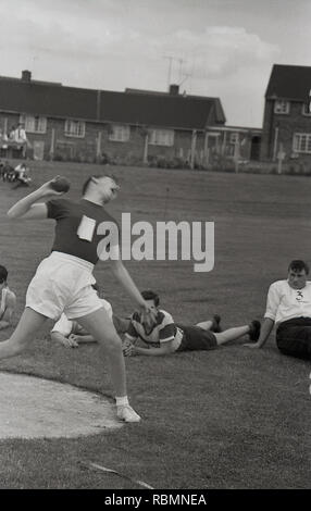 1960er Jahre, historisch, Schulsport Tag, Schuljunge in Shorts macht eine große Anstrengung, als er versucht, werfen oder schieben die "heiß", das schwere Gewicht in der Schuss Put-Veranstaltung, Englabd, Großbritannien. Der Kugelschuss ist eine fantastische Sportart, die sowohl gute Technik als auch Kraft aufzeigt und seit 1896 in den Olympischen Spielen der Moderne zu sehen ist. Seine Ursprünge liegen angeblich beim Wettbewerb „Putting the Stone“ bei den traditionellen Scottish Highland Games. Stockfoto