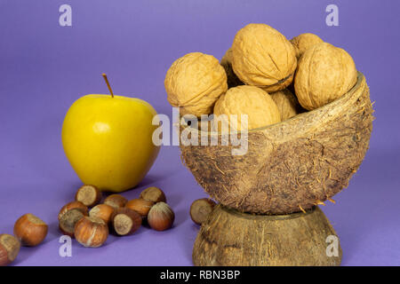 Gesamten gemeinsamen Walnüsse mit Schale in natürlichen Coconut shell Cup, Haufen von ganzen Haselnüssen und gelber Apfel auf lila Hintergrund Stockfoto