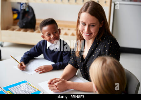 Ansicht eines weiblichen Lehrer zwischen zwei Schulkinder am Tisch in einem Klassenzimmer sitzen, an jedem anderen lächelnd während einer Lektion Stockfoto