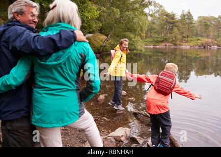 Zwei Kinder spielen am Ufer eines Sees, ihre Großeltern im Vordergrund, Lake District, Großbritannien Stockfoto