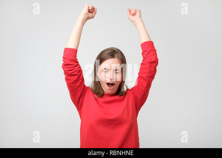 Glückliche junge Frau in Rot tragen Gestik und halten die Augen beim Stehen vor grauem Hintergrund geschlossen. Erfolge feiern. Stockfoto