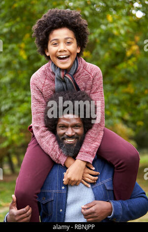 Schwarz im mittleren Alter Mann seinen Sohn auf seinen Schultern in den Park, die beide lächeln in die Kamera, in der Nähe Stockfoto